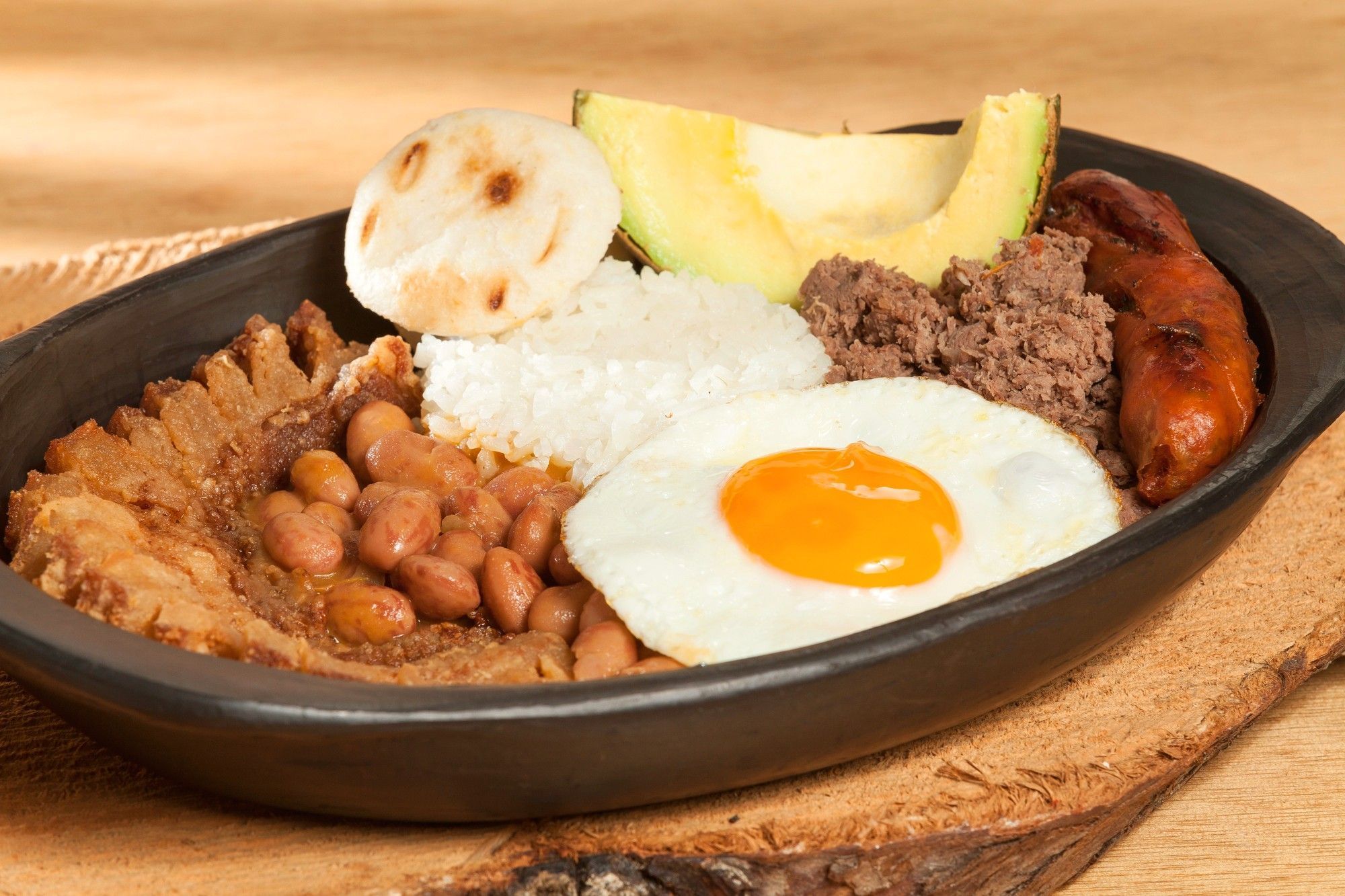 A bandeja paisa in a black clay dish: chicharrón, beans, rice, fried egg, ground beef, chorizo, arepa and avocado