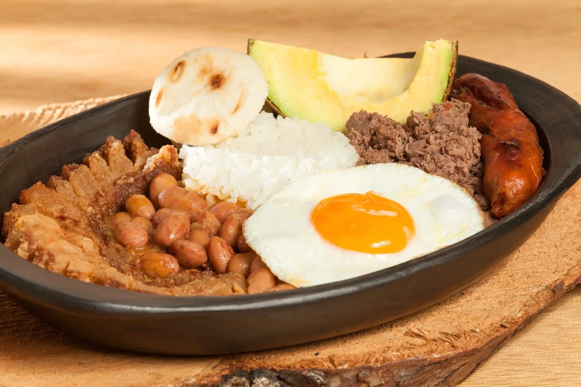 A bandeja paisa in a black clay dish: chicharrón, beans, rice, fried egg, ground beef, chorizo, arepa and avocado