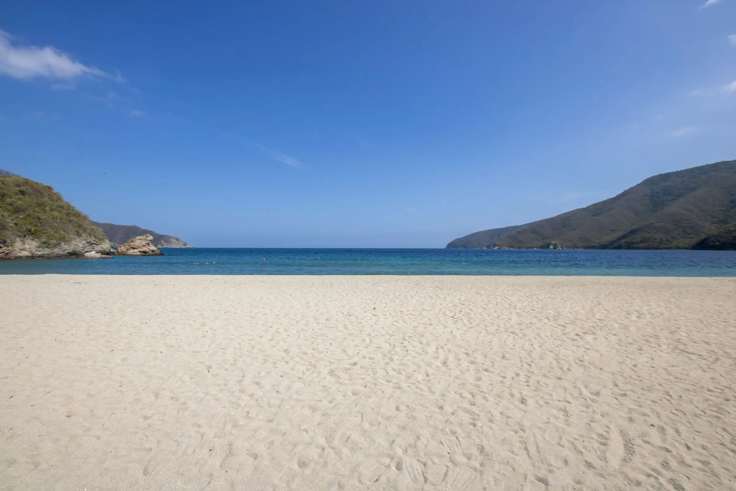 Una playa de arena clara en el mar Caribe, con cerros verdes a los lados y el cielo despejado
