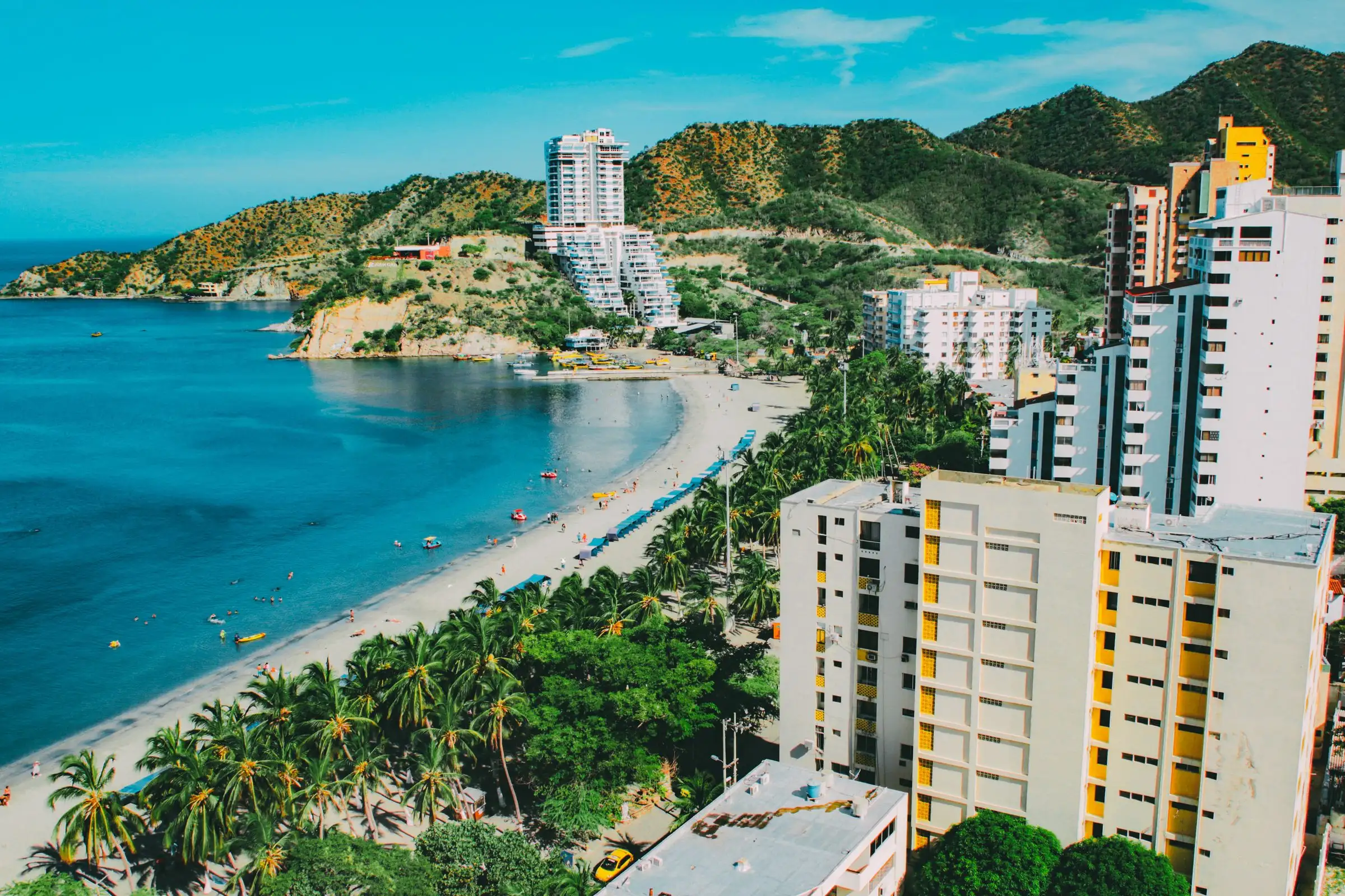 A beach in Santa Marta with palm trees, seafront buildings and green hills in the background